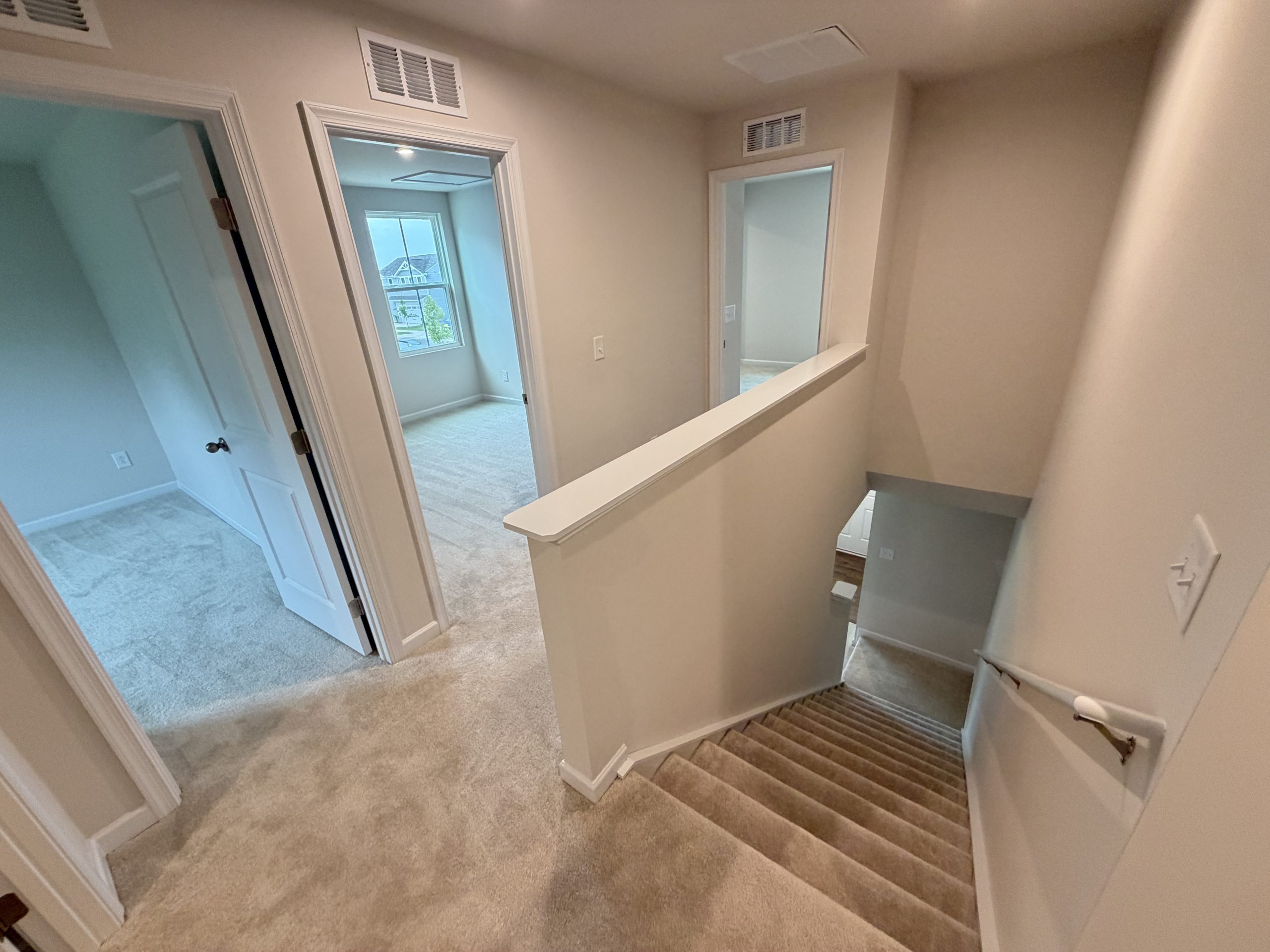 Interior stair landing with carpeted stairs descending on the right, beige walls, white trim, and doorways to rooms; window shows houses outside in the distance.