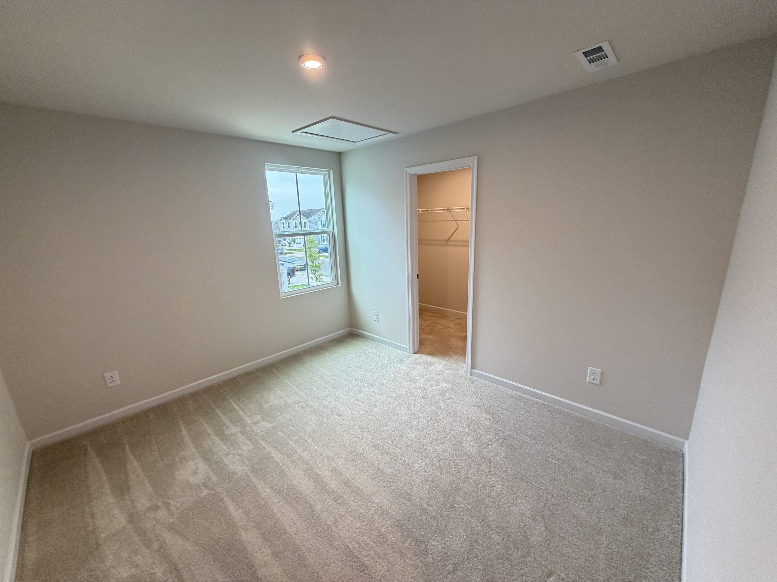 Empty beige bedroom with a window, closet door ajar, and ceiling light fixture; carpeted floor.