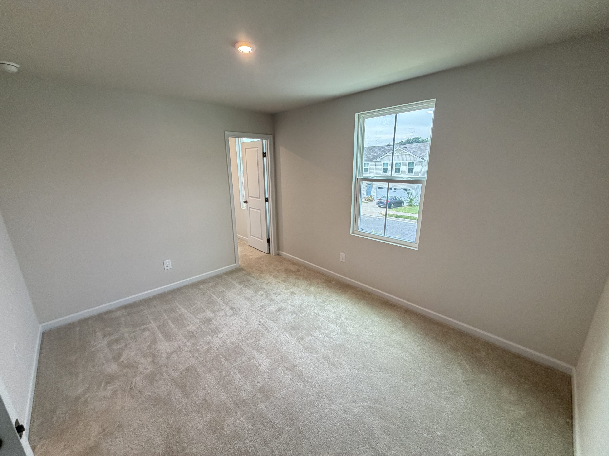 Small empty bedroom with beige carpet, light gray walls, a window overlooking a suburban street, and an open doorway.