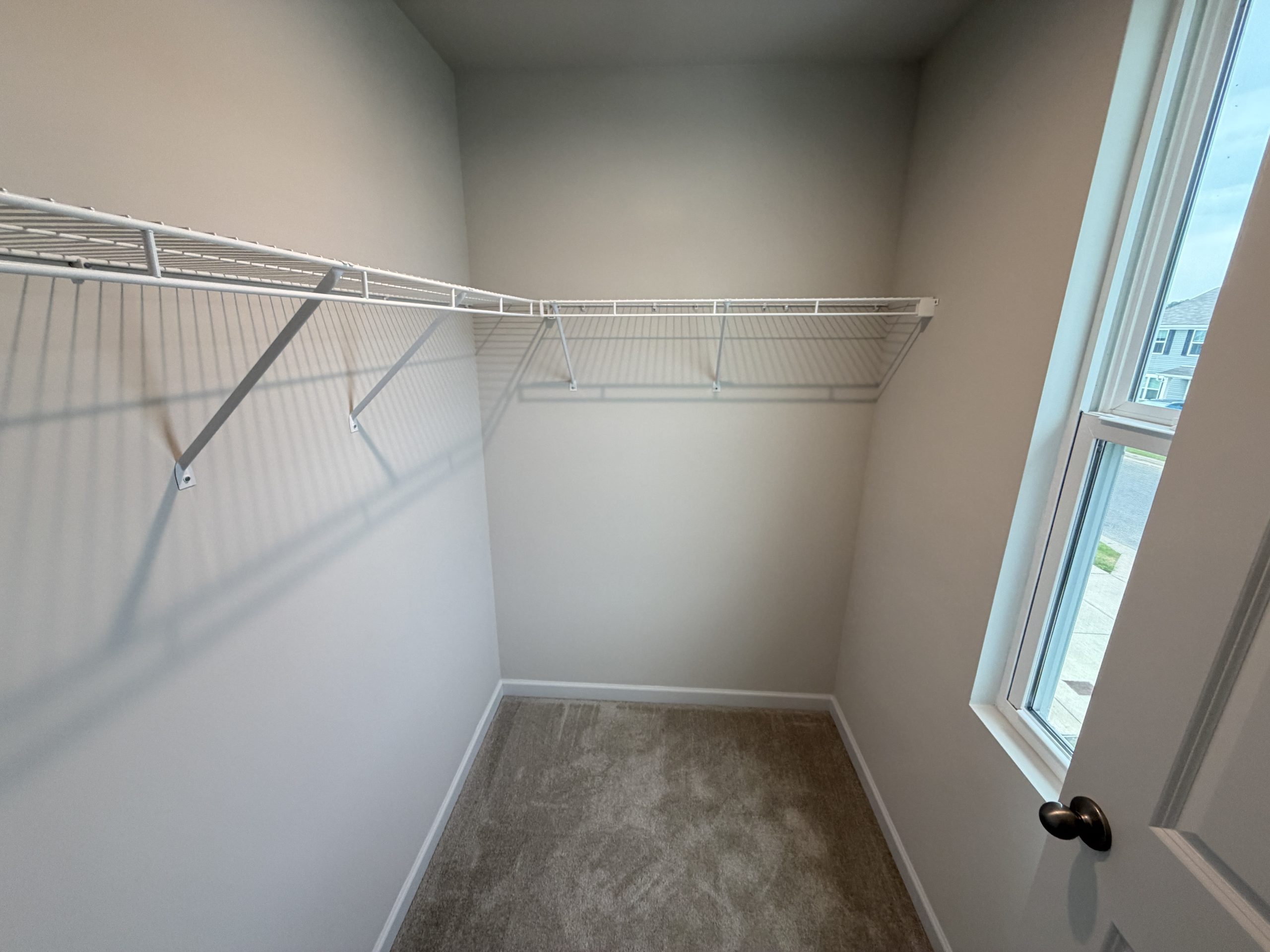 Empty walk-in closet with white wire shelving along two walls and daylight coming from a large window on the right side.
