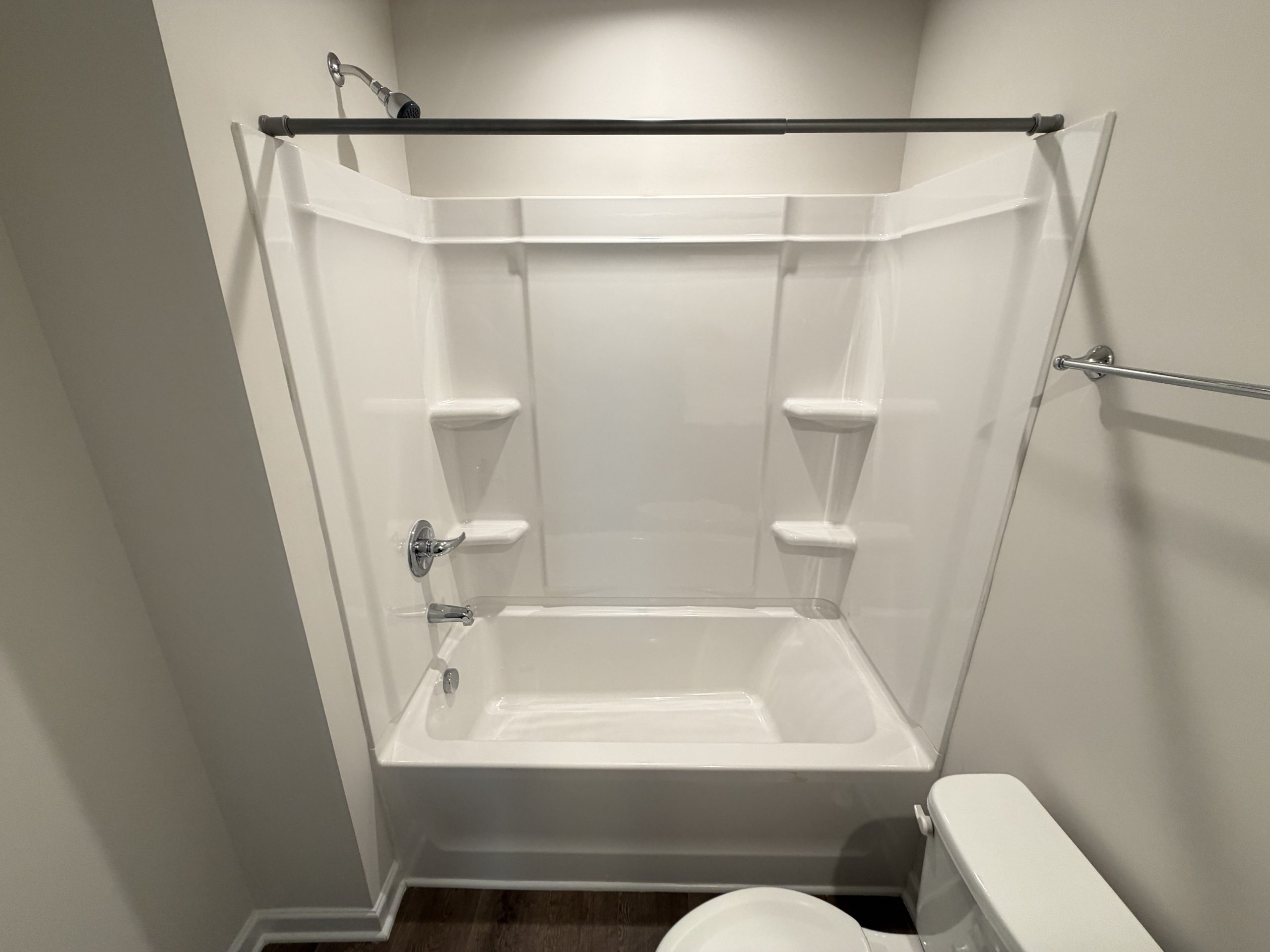 White alcove shower/tub with built-in seating and a horizontal shower rod across the top; bathroom with a nearby toilet visible on the right.