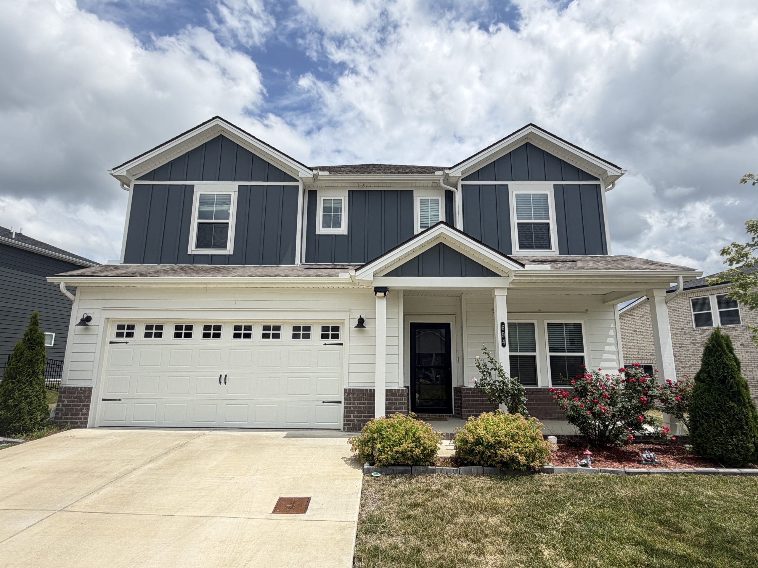 Two-story blue suburban house with a white garage door and a covered entry, blue siding, and a manicured front yard under a partly cloudy sky