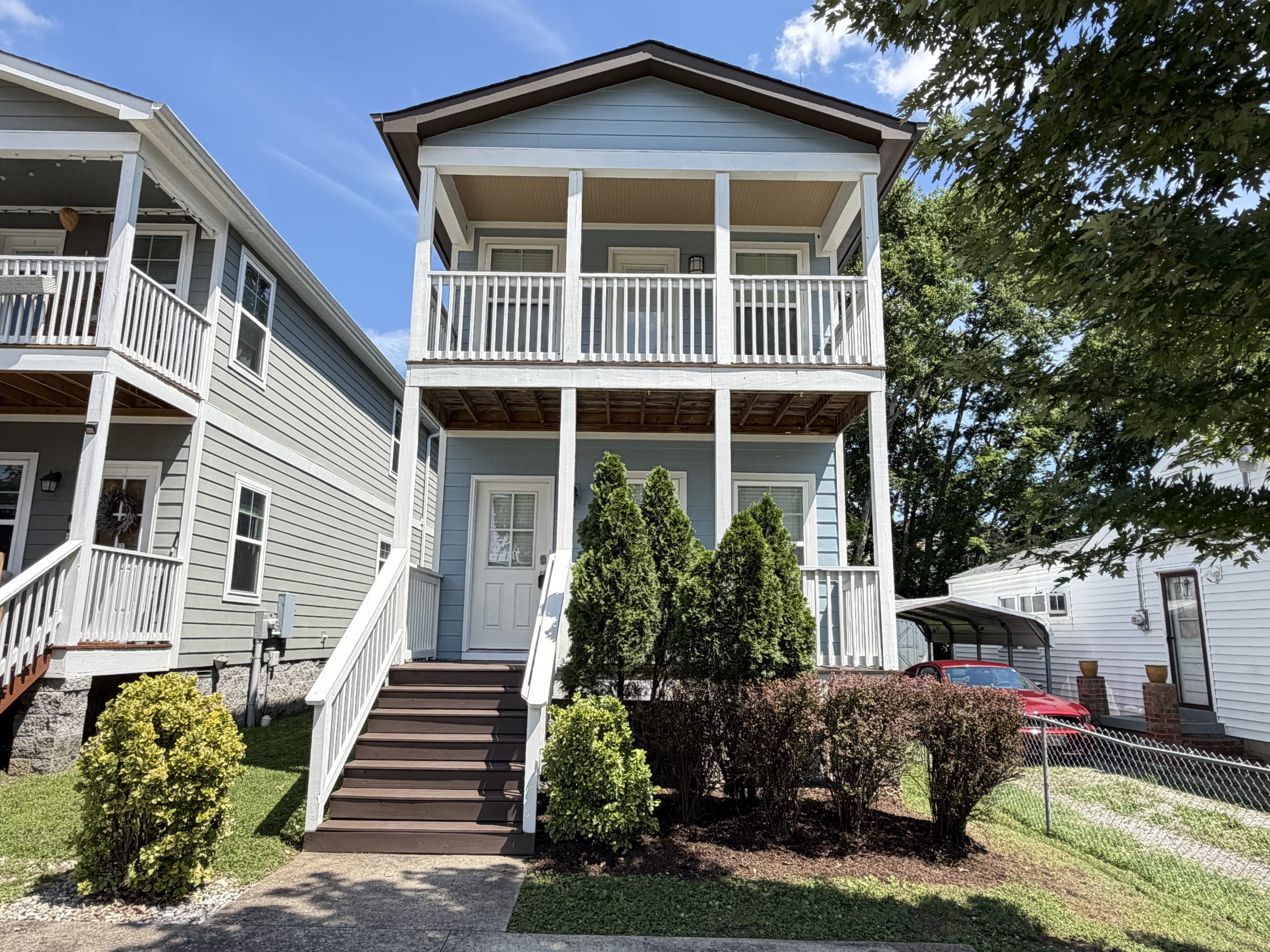 Two-story blue townhouse with a white railing porch and stairs, front door, and manicured shrubs under a clear sky
