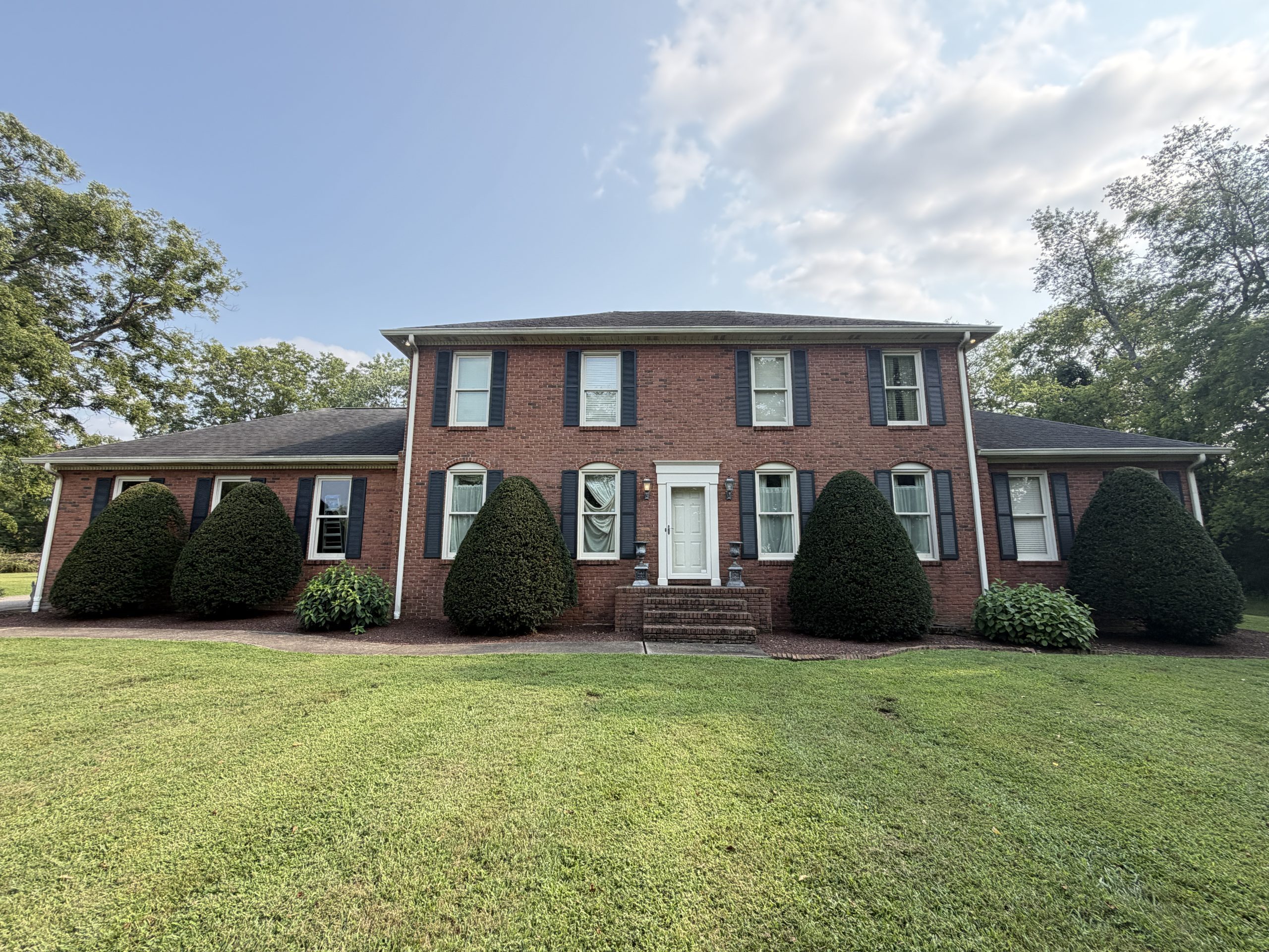 Front view of a brick two-story house with white trim, black shutters, and a row of rounded evergreen shrubs in the Yard under a blue sky.