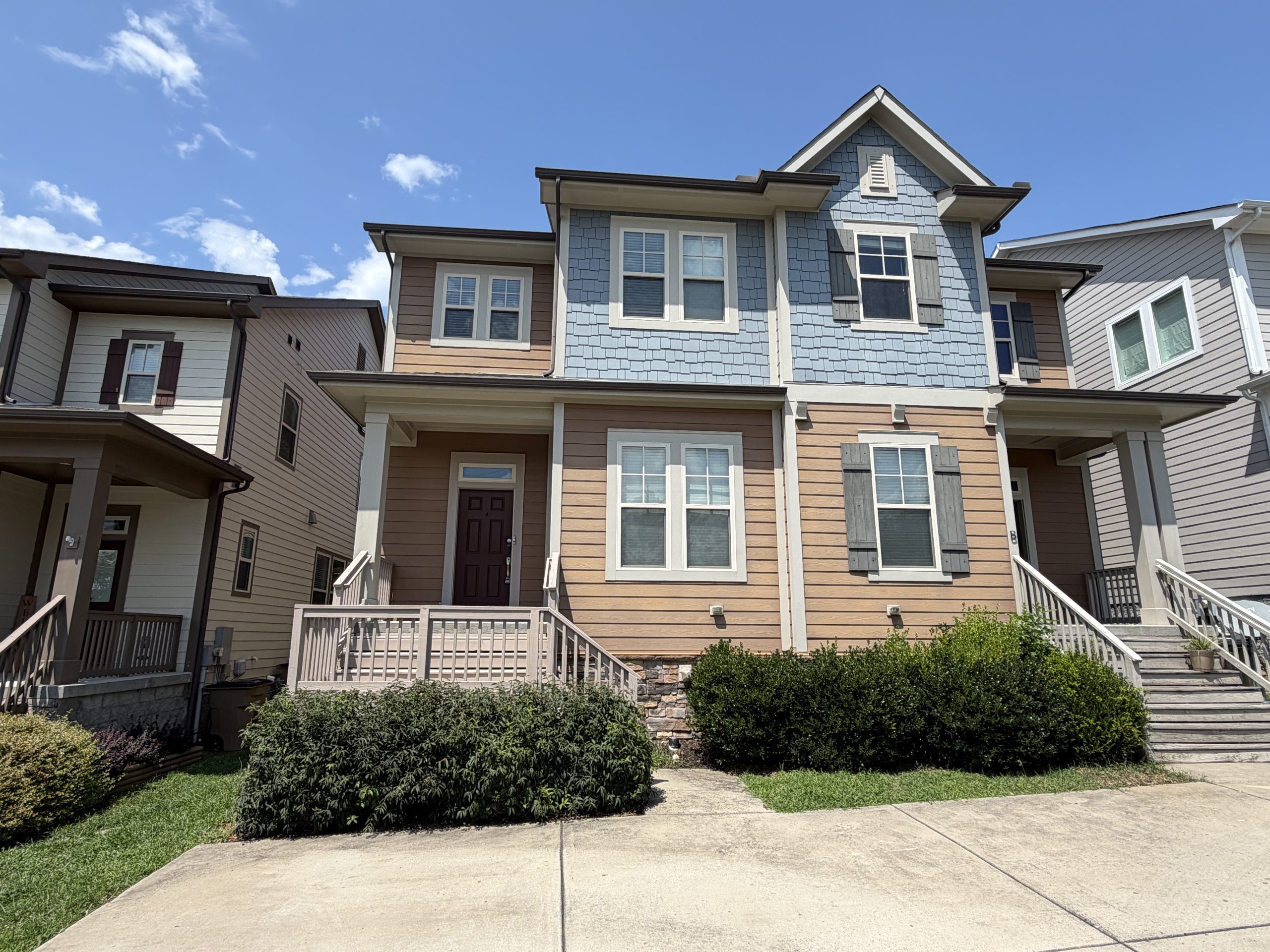Front view of a row of beige and blue-gray townhouses with stairs, shrubs, and a bright blue sky above.